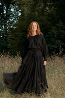 Woman in a long black dress standing in a field with trees in the background