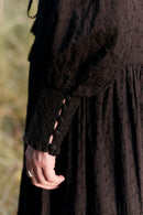 Close-up of a person wearing a dark brown dress with lace sleeves against a blurred natural background.