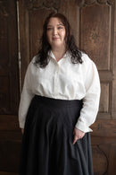 Woman wearing a white blouse and dark skirt standing against a wooden paneled wall.