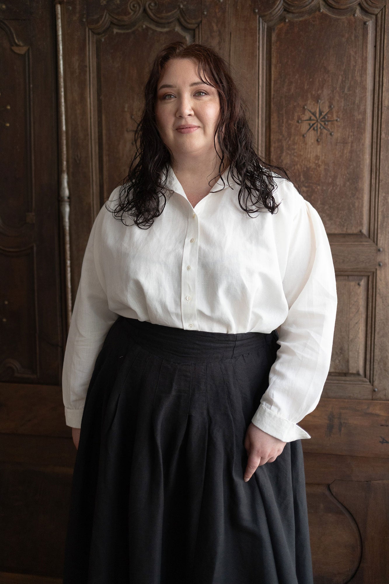 Woman wearing a white blouse and dark skirt standing against a wooden paneled wall.