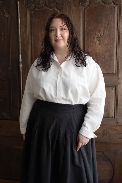 Woman wearing a white blouse and dark skirt standing against a wooden paneled wall.