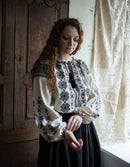 Woman wearing a traditional embroidered blouse in a room with wooden paneling and lace curtains.
