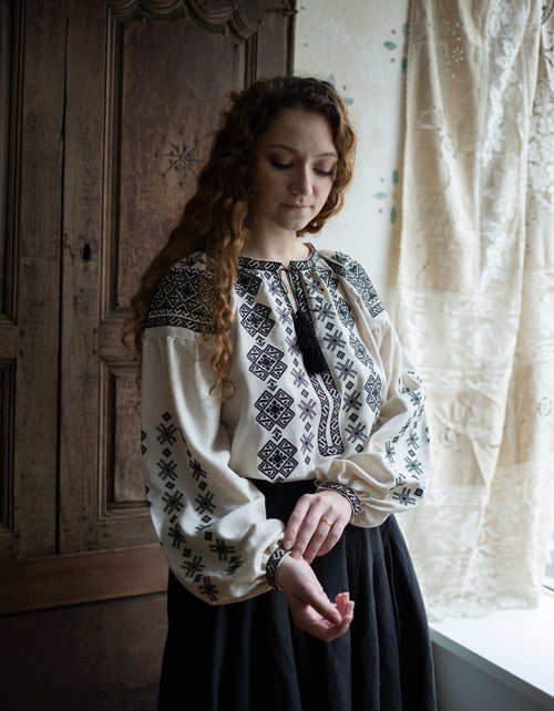 Woman wearing a traditional embroidered blouse in a room with wooden paneling and lace curtains.