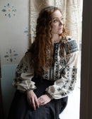 Woman wearing a traditional embroidered blouse sitting by a window.