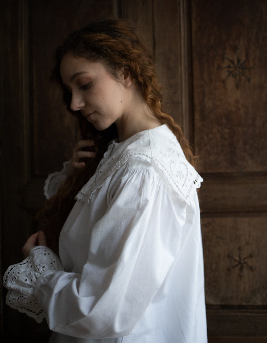 Woman wearing a white blouse with lace details in a dimly lit room.