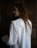 Woman wearing a white blouse with lace details in a dimly lit room.