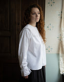 Woman wearing a white blouse with lace cuffs in a room with wooden paneling and floral wallpaper.