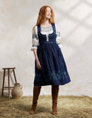 Woman wearing a traditional embroidered dress with a white blouse and brown boots, standing on hay with a rustic background.