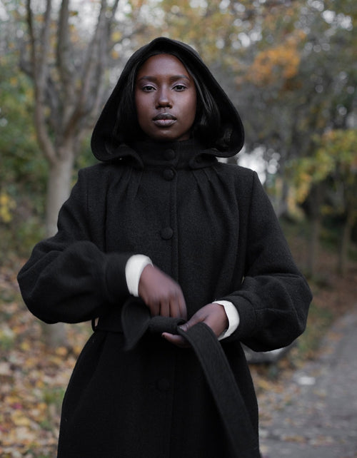 Person wearing a black coat with a hood in an outdoor setting with trees and leaves.