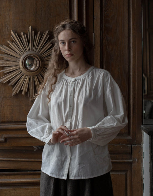 Woman wearing a white blouse standing in front of a wooden wall with decorative elements.