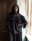 Woman wearing a dark ruffled blouse and skirt in a room with wooden paneling and a window.