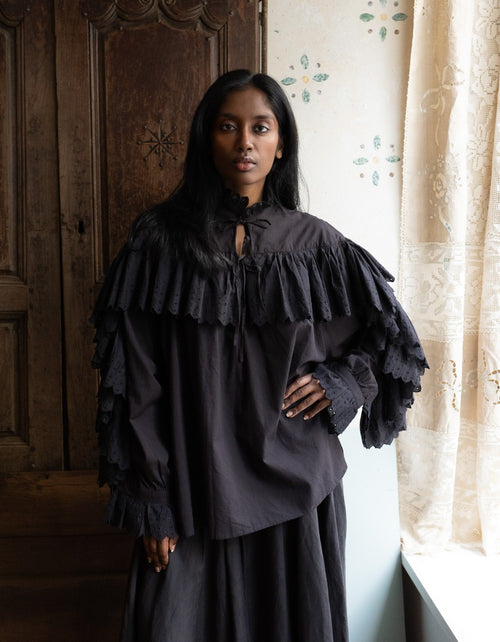Woman wearing a dark ruffled blouse and skirt in a room with wooden paneling and a window.