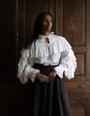 Woman wearing a white blouse with ruffled sleeves and a black skirt against a wooden paneled wall.