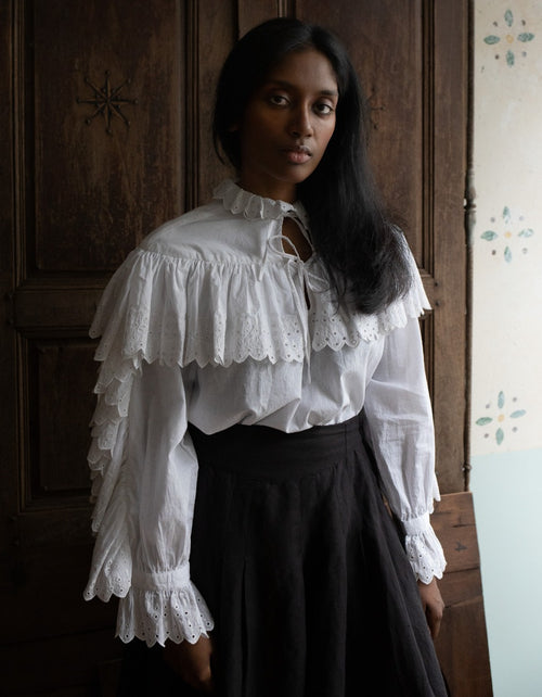 Woman wearing a white blouse with ruffled sleeves and black skirt in a room with wooden paneling.