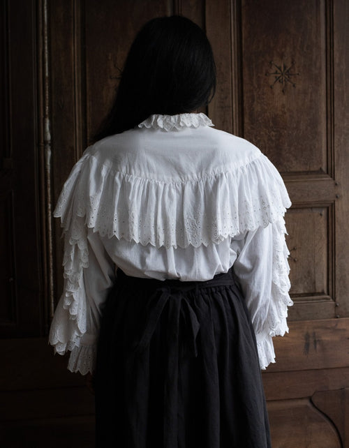 Person wearing a white blouse with ruffled details in front of a wooden door.