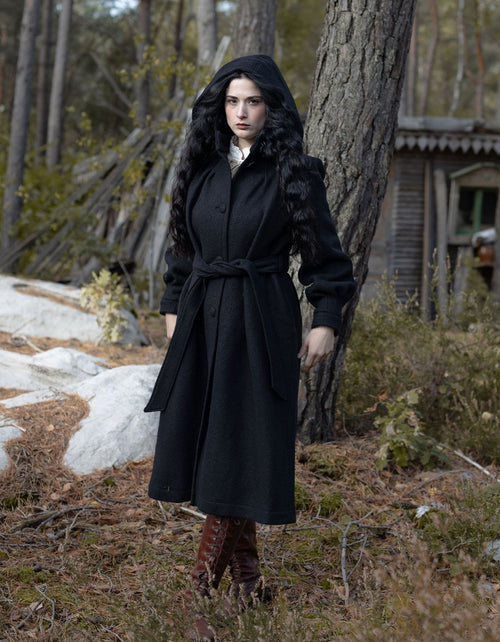 Woman in a black coat standing in a forested area with a rustic building in the background.