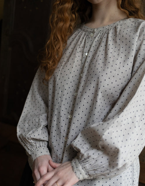 Person wearing a light-colored blouse with a subtle pattern, standing against a dark background.