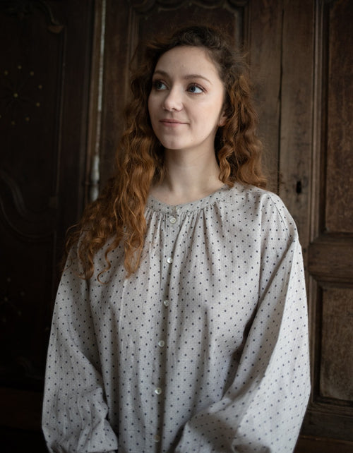 Woman wearing a light blouse with a polka dot pattern in a dimly lit room.