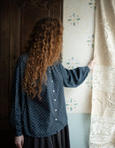 Woman in a polka dot blouse standing next to a curtain with floral patterns.