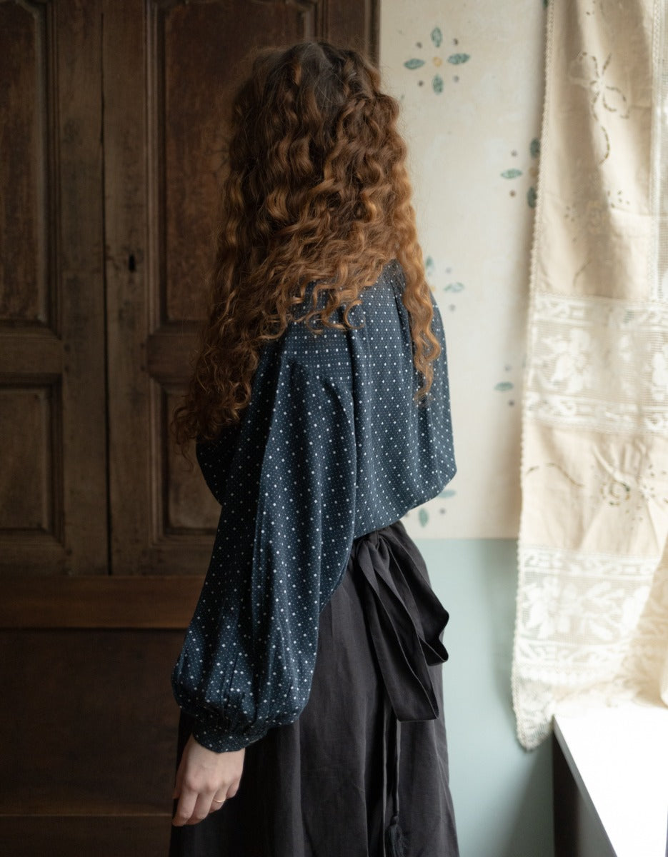 Person wearing a dark blue polka dot blouse with long hair, standing in a room with wooden paneling and a curtain.