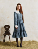 Woman wearing a blue dress with white lace details standing next to a wooden chair on hay.