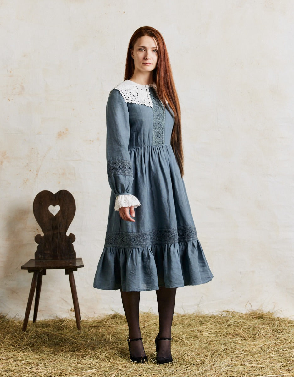 Woman wearing a blue dress with white lace details standing next to a wooden chair on hay.