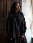 Woman wearing a black long-sleeve top with lace details in a room with wooden paneling and a window.