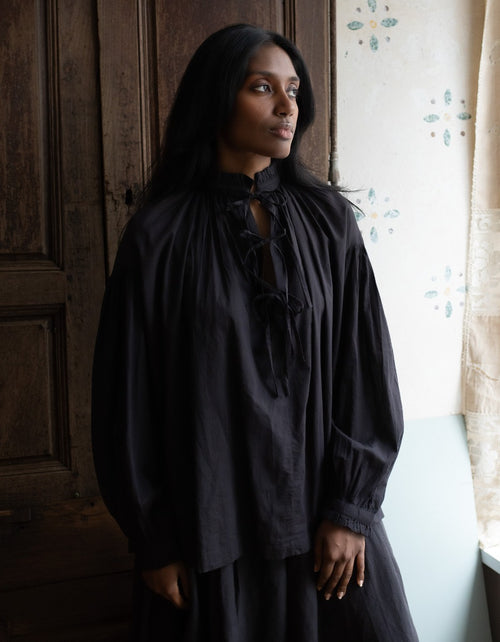 Woman wearing a black long-sleeve top with lace details in a room with wooden paneling and a window.