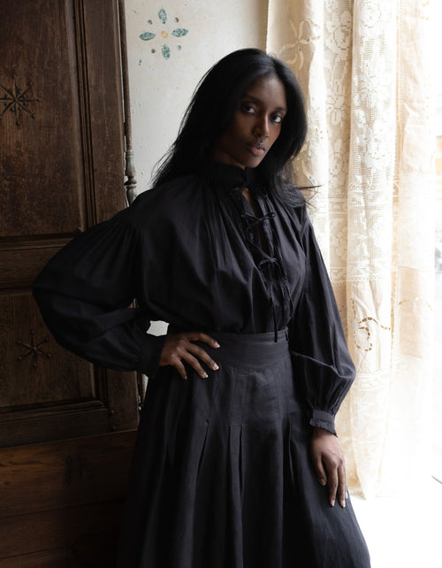 Woman wearing a dark dress standing in a room with wooden paneling and curtains.