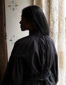 Person wearing a black dress standing in a softly lit room with curtains.