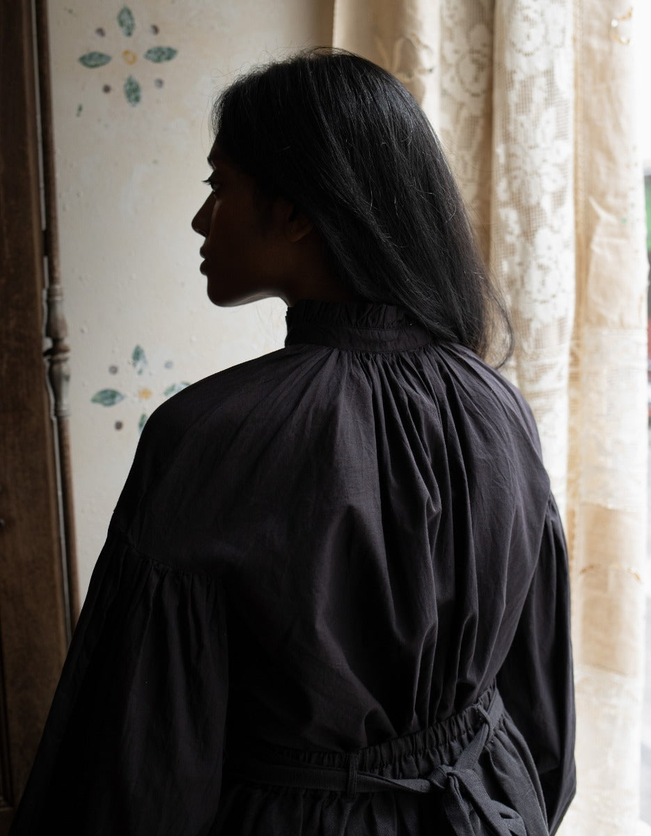 Person wearing a black dress standing in a softly lit room with curtains.