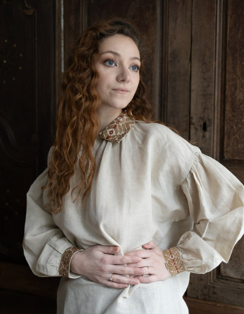 Woman in a historical or period-style dress with intricate details, standing against a wooden paneled wall.