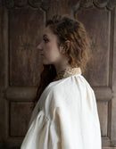 Woman in a white garment with a textured collar against a wooden paneled wall.