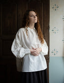 Woman wearing a white blouse standing in a room with wooden walls and floral wallpaper.
