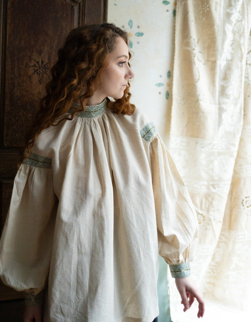 Woman wearing a historical-style dress with intricate embroidery in a softly lit room.