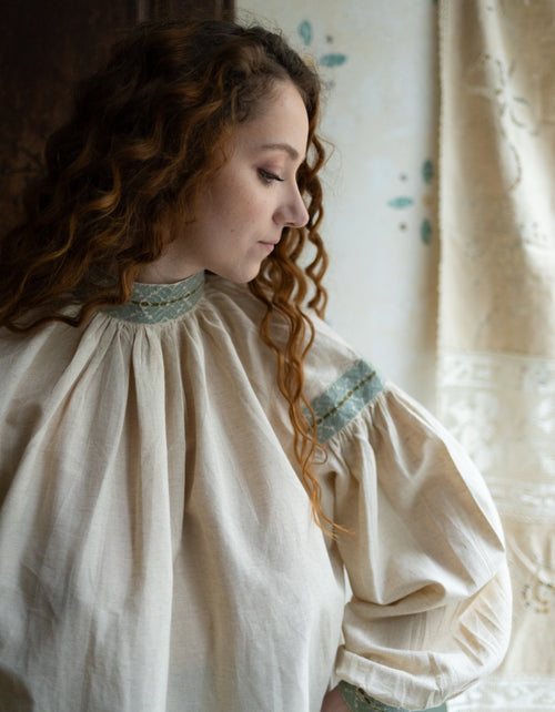 Woman wearing a beige blouse with lace details in a softly lit room.