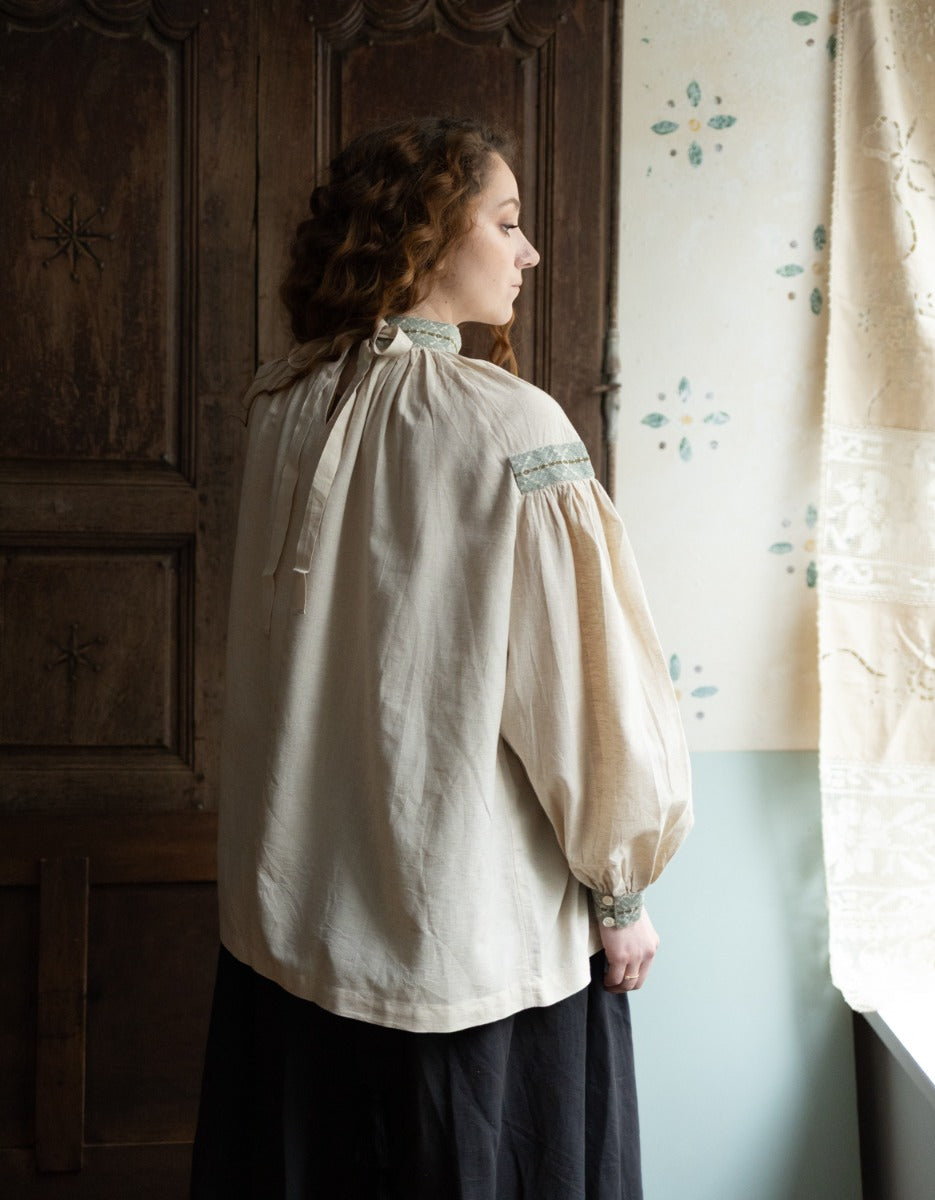 Woman wearing a beige blouse with embroidered details, standing in a room with wooden paneling and floral curtains.