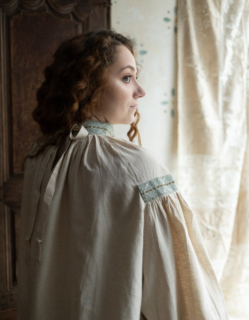 Woman in a historical-style dress standing in a room with curtains.