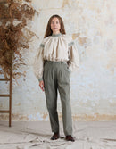 Woman wearing a beige blouse with green accents and gray pants against a textured wall.